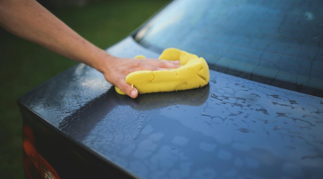 Hand cleaning car with a yellow sponge, showcasing car detailing and washing process.