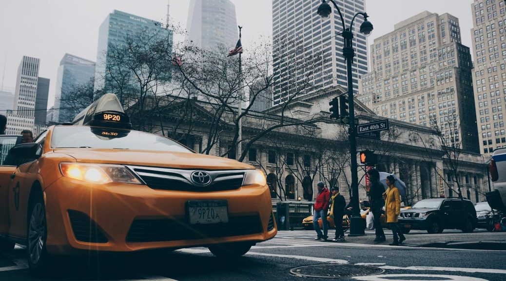 A bustling New York City street intersection featuring a yellow taxi and pedestrians with skyscrapers in the background.