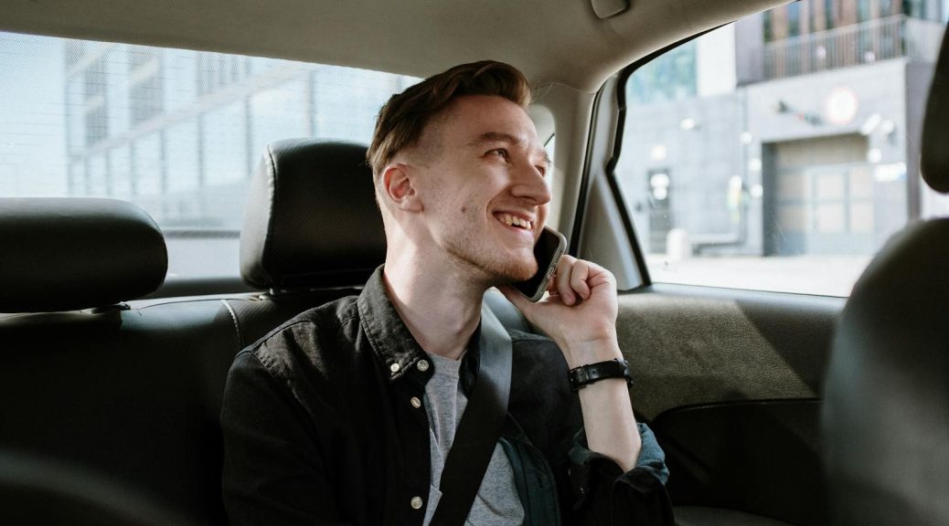 Smiling young man enjoying a phone call while seated in the back seat of a car during a daytime ride.
